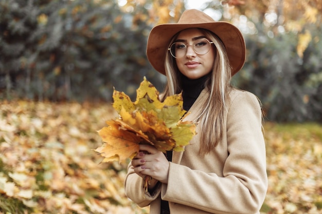 Jeune femme drôle vêtue d'un manteau et d'un chapeau tient une brassée de feuilles mortes dans le parc de l'automne