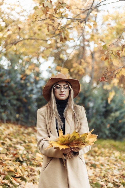 Jeune femme drôle vêtue d'un manteau et d'un chapeau tient une brassée de feuilles mortes dans le parc de l'automne