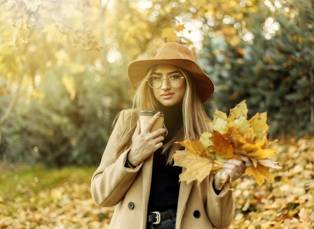 Jeune femme drôle vêtue d'un manteau et d'un chapeau tient une brassée de feuilles tombées dans le parc en automne. Plaisir d'automne