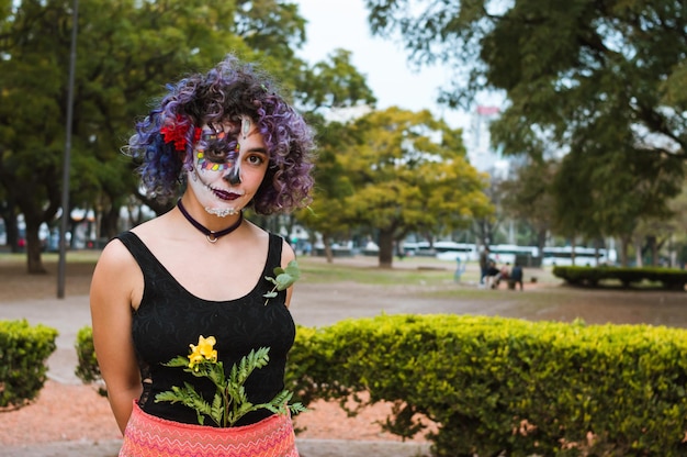 Jeune femme avec du maquillage de catrina et des vêtements folkloriques se tient dans le parc en regardant la caméra