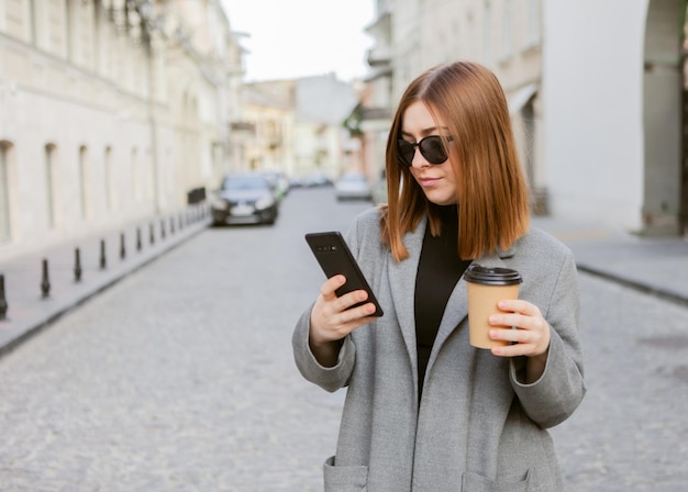 Une jeune femme du millénaire aux cheveux lisses vêtue d'un manteau d'automne et de lunettes de soleil utilise un smartphone et tient une tasse de café en déplacement dans une ville européenne.