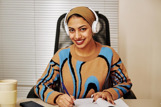 Jeune femme du Moyen-Orient portant des écouteurs au bureau et souriant à la caméra