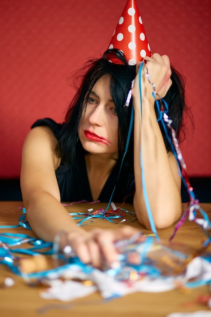 Jeune femme avec du rouge à lèvres et une casquette frottés rouges, assise fatiguée à table dans une pièce en désordre après la fête d'anniversaire