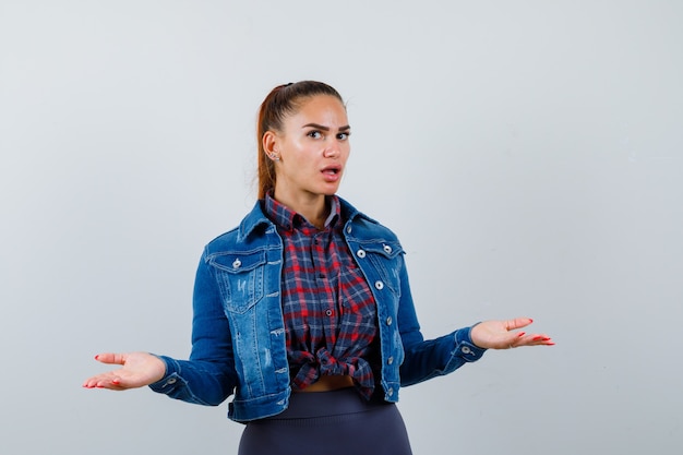 Jeune femme écartant les bras en chemise à carreaux, veste en jean et l'air surpris, vue de face.