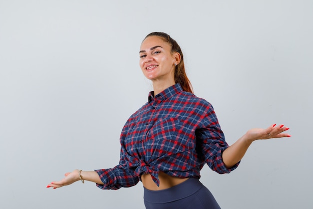 La jeune femme écarte les paumes en chemise à carreaux, en pantalon et a l'air heureuse. vue de face.