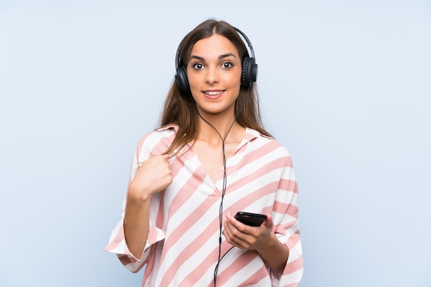 Jeune femme écoutant de la musique avec un mur bleu isolé mobile avec une expression faciale surprise