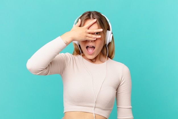 Jeune femme avec des écouteurs ayant l'air choquée, effrayée ou terrifiée, couvrant le visage avec la main