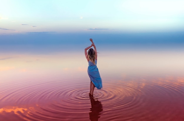 Photo jeune femme élancée attrayante dans une robe bleue dans l'eau rose d'un lac salé, sur une surface de ciel bleu