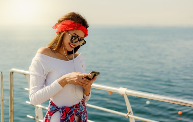 Une jeune femme élégamment vêtue écoute de la musique dans des écouteurs et utilise un smartphone sur la plage sur fond de mer