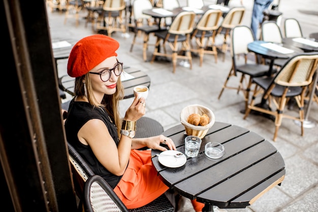 Photo jeune femme élégante en béret rouge prenant un petit-déjeuner français avec café et croissant assis à l'extérieur à la terrasse du café