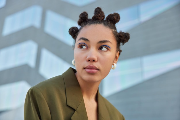 Une jeune femme élégante avec des chignons concentrés avec une expression sérieuse porte un eye-liner bleu vif vêtu d'une veste élégante verte pose sur fond flou. Concept de mode de vie de rue