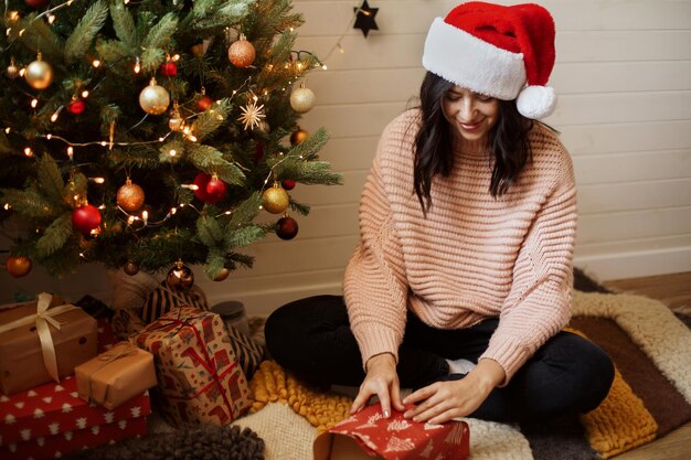 Jeune femme élégante enveloppant le cadeau de Noël dans du papier sous le sapin de Noël avec des lumières dans une chambre moderne