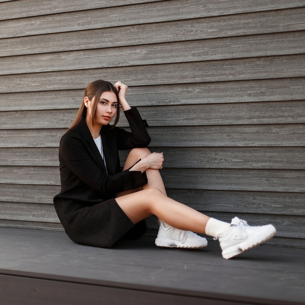 Jeune femme élégante avec des jambes sexy dans un manteau noir avec des baskets blanches assis près d'un mur en bois