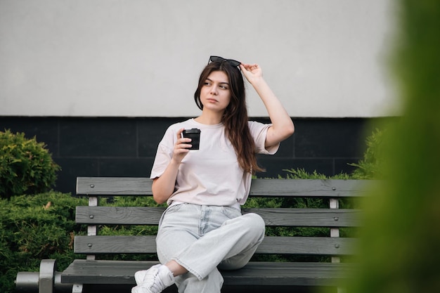 Une jeune femme élégante en lunettes de soleil est assise sur un banc et boit du café