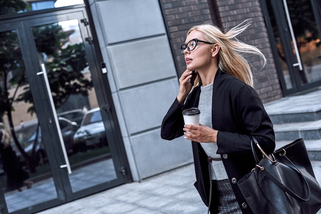 Jeune femme élégante parlant par smartphone, elle marche avec le café du matin