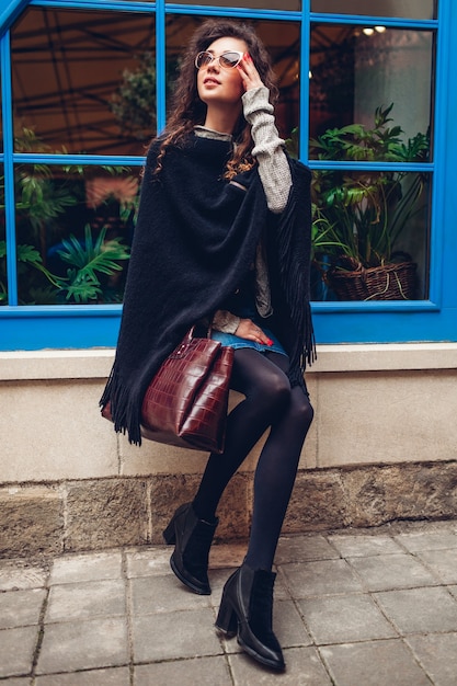 Jeune femme élégante posant contre la fenêtre bleue à l'extérieur. Tenue à la mode. Beau modèle aux cheveux bouclés souriant