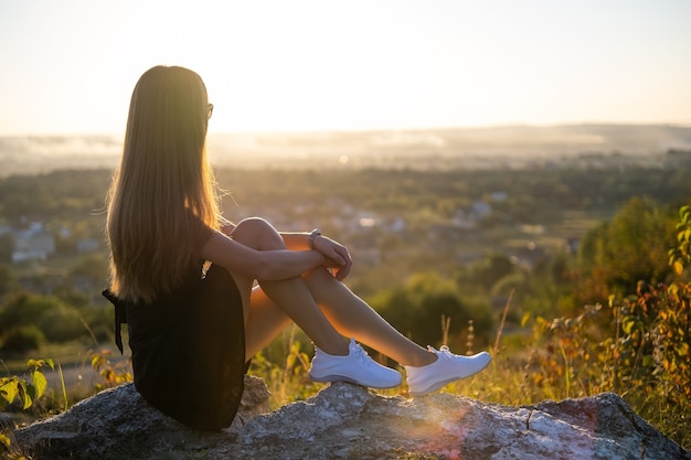 Jeune femme élégante en robe courte noire et chaussures baskets blanches assises sur un rocher relaxant à l'extérieur le soir d'été. Dame à la mode profitant d'un coucher de soleil chaud dans la nature.