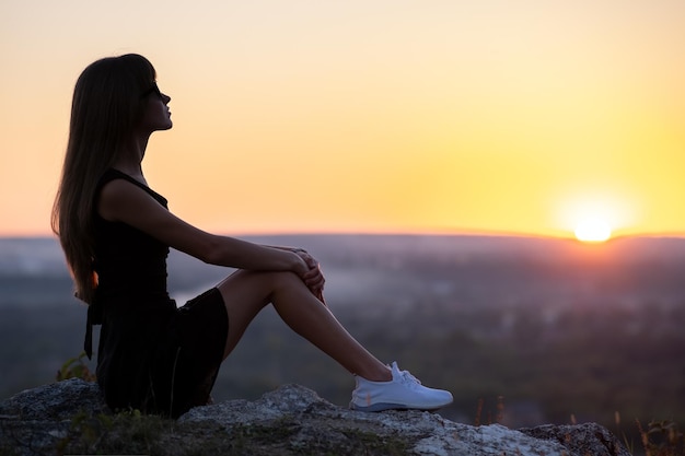 Jeune femme élégante en robe courte noire et chaussures baskets blanches assises sur un rocher relaxant à l'extérieur le soir d'été. Dame à la mode profitant d'un coucher de soleil chaud dans la nature.