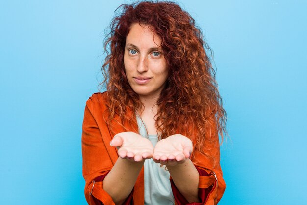 Photo jeune femme élégante rousse tenant quelque chose avec des paumes, offrant à la caméra.