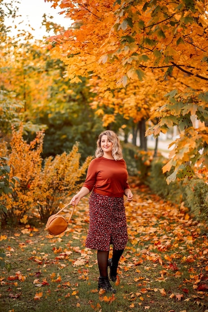 Jeune femme élégante de taille plus avec un bouquet de feuilles jaunes se promène dans un parc d'automne Belle fille souriante dans un costume marron