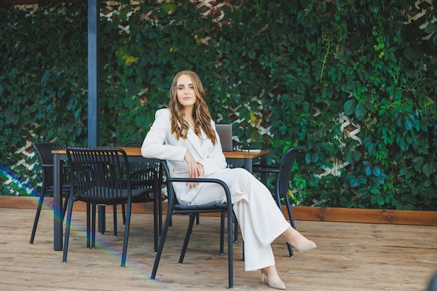 Une jeune femme élégante vêtue d'un costume classique blanc avec un sourire sur son visage se tient sur la terrasse d'un café d'été