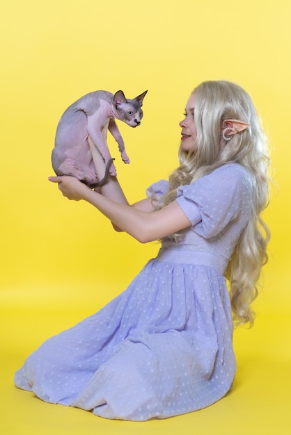 Jeune femme elfe cosplay assise sur fond jaune montre un chaton sphinx le tenant dans les mains