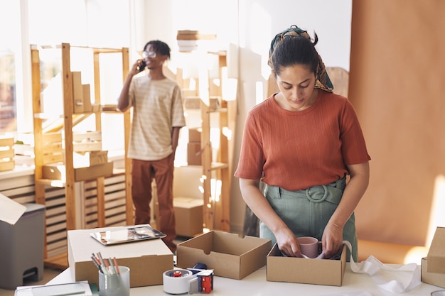 Jeune femme d'emballage des colis dans les boîtes à la table avec son collègue parlant au téléphone mobile en arrière-plan, ils travaillent dans le service de livraison