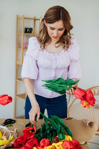 Une jeune femme emballe un bouquet festif dans du papier d'emballage Le fleuriste fait un assemblage avec des tulipes rouges à l'atelier Une femme au travail dans une petite entreprise ou passe-temps