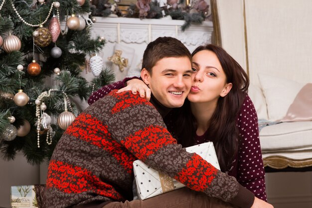 Jeune femme embrassant son petit ami pour le remercier de son cadeau de Noël alors qu'ils sont assis par terre devant l'arbre de Noël