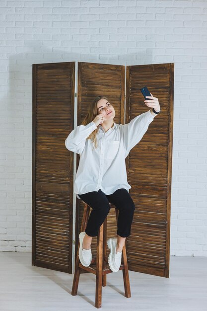 Une jeune femme émotionnelle dans une chemise en coton blanc gesticule et regarde l'écran d'un gadget moderne Une fille avec un téléphone portable prend un selfie