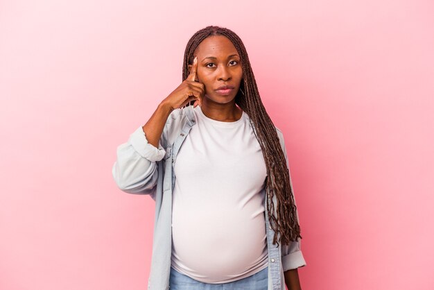 Jeune femme enceinte afro-américaine isolée sur fond rose pointant le temple avec le doigt, pensant, concentrée sur une tâche.