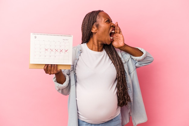 Jeune femme enceinte afro-américaine tenant un calendrier isolé sur fond rose criant et tenant la paume près de la bouche ouverte.