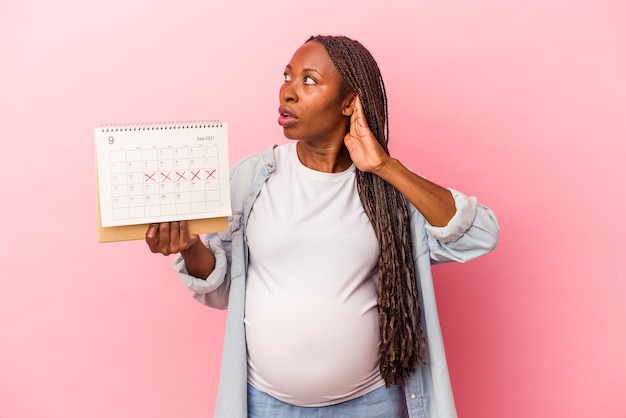 Jeune femme enceinte afro-américaine tenant un calendrier isolé sur fond rose essayant d'écouter un potin.