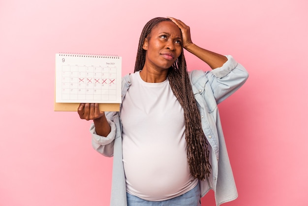 Jeune Femme Enceinte Afro-américaine Tenant Un Calendrier Isolé Sur Fond Rose étant Choquée, Elle S'est Souvenue D'une Réunion Importante.