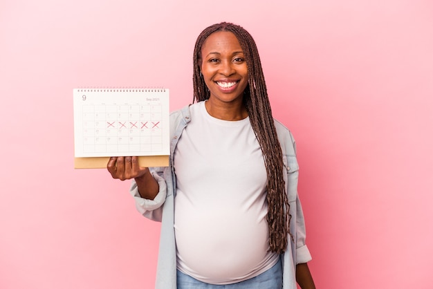 Jeune femme enceinte afro-américaine tenant un calendrier isolé sur fond rose heureux, souriant et joyeux.