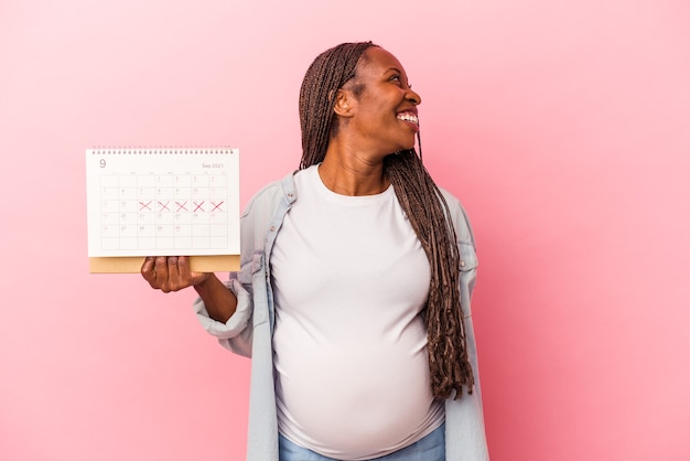 Photo jeune femme enceinte afro-américaine tenant un calendrier isolé sur fond rose regarde de côté souriant, joyeux et agréable.