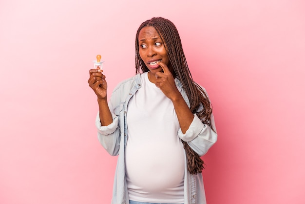 Jeune femme enceinte afro-américaine tenant une tétine isolée sur fond rose détendue en pensant à quelque chose en regardant un espace de copie.