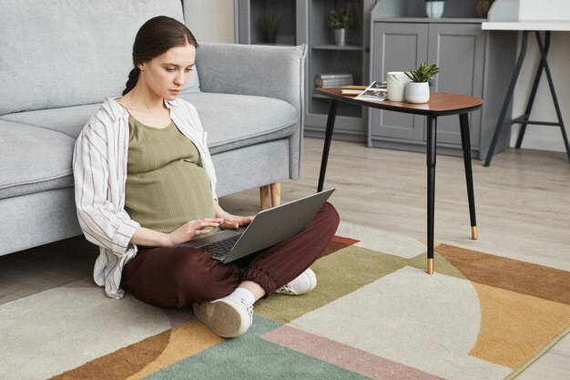 Jeune femme enceinte assise sur le sol avec un ordinateur portable sur les genoux et travaillant en ligne dans la chambre à la maison