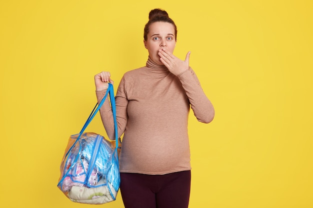 Jeune Femme Enceinte Attrayante Portant Avec Désinvolture, Tenant Un Sac Avec Des Trucs, étonnée Et Peur D'aller à La Maternité, Couvrant La Bouche Avec La Paume.