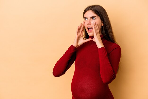 Jeune femme enceinte caucasienne isolée sur fond beige crie fort, garde les yeux ouverts et les mains tendues.