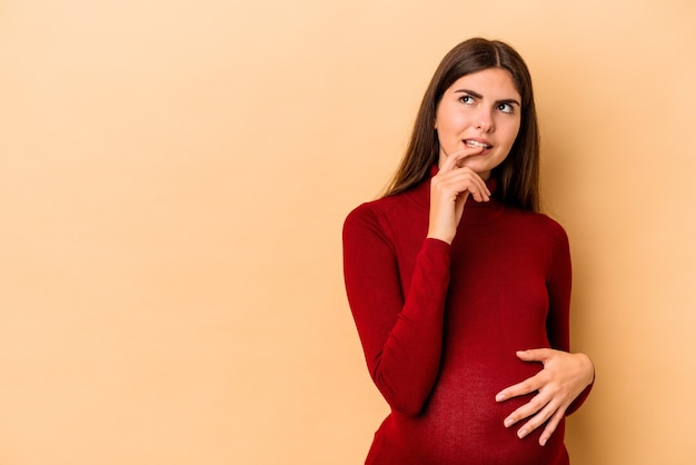 Jeune femme enceinte caucasienne isolée sur fond beige détendue en pensant à quelque chose en regardant un espace de copie.