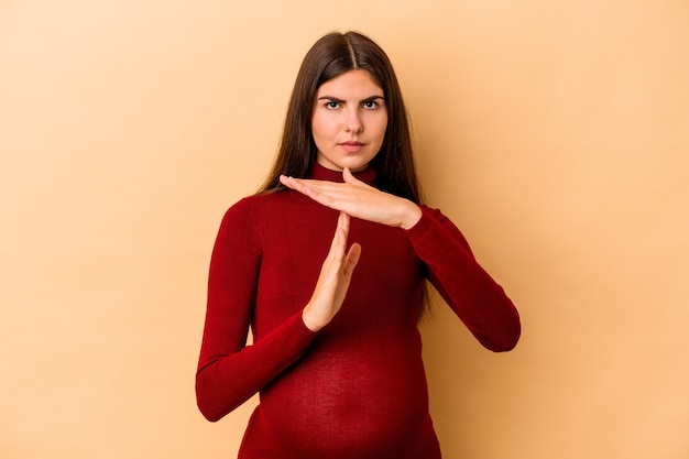 Jeune femme enceinte caucasienne isolée sur fond beige montrant un geste de délai d'attente.