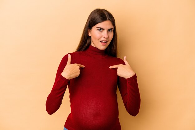 Jeune femme enceinte caucasienne isolée sur fond beige surpris pointant avec le doigt, souriant largement.