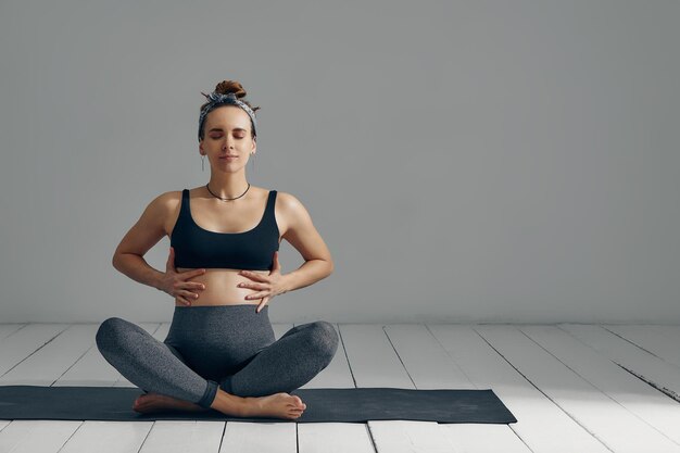 Une jeune femme enceinte du millénaire assise sur le tapis touche son ventre après avoir effectué des exercices prénataux et de méditation lors d'un cours de yoga Concept de vie et de maternité