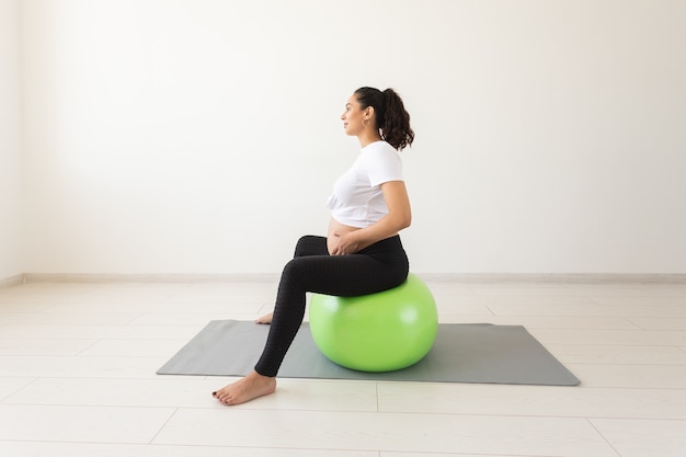 Une jeune femme enceinte faisant des exercices de relaxation à l'aide d'un ballon de fitness assis sur un tapis et tenant son ventre