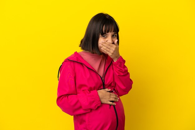 Photo jeune femme enceinte sur fond jaune isolé heureux et souriant couvrant la bouche avec la main