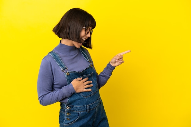 Jeune femme enceinte sur fond jaune isolé, pointant le doigt sur le côté et présentant un produit