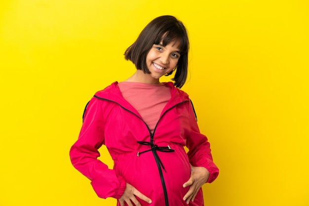 Jeune femme enceinte sur fond jaune isolé posant avec les bras à la hanche et souriant