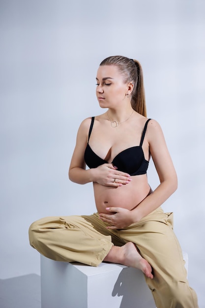 Jeune femme enceinte heureuse en soutien-gorge et pantalon sur fond blanc Grossesse heureuse Femme enceinte élégante pose dans le studio