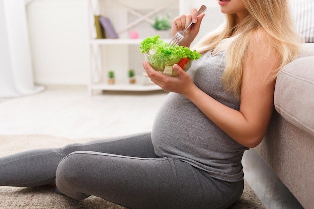 Jeune femme enceinte mangeant une salade verte en gros plan. Blonde enceinte méconnaissable assise par terre et prenant une collation fraîche. Concept de nutrition saine et de grossesse